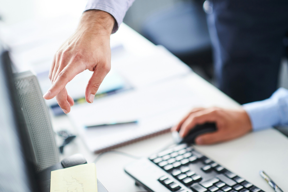 Two colleagues working side by side at an office desk, one reaching towards a monitor